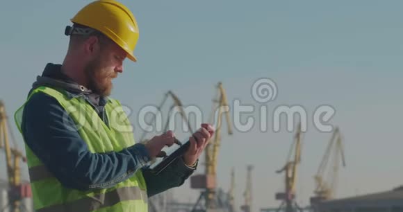 港口工人与胡子在一个黄色头盔的立场与平板电脑在海港的背景起重机领班视频的预览图