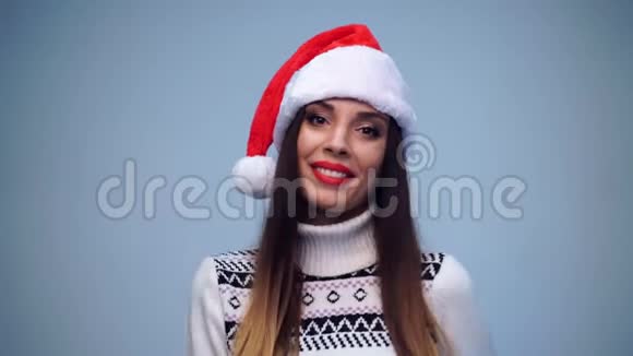 年轻漂亮的黑发女郎穿着白色毛衣和圣诞老人的帽子站在蓝底的工作室里微笑着看着视频的预览图