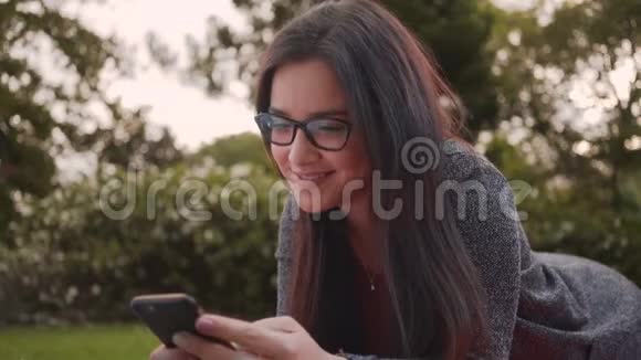 一位迷人的微笑的年轻女子躺在公园里在智能手机上发短信的画像视频的预览图