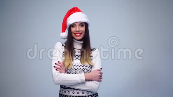 年轻漂亮的黑发女郎穿着白色毛衣和圣诞老人的帽子站在蓝底的工作室里微笑着看着视频的预览图
