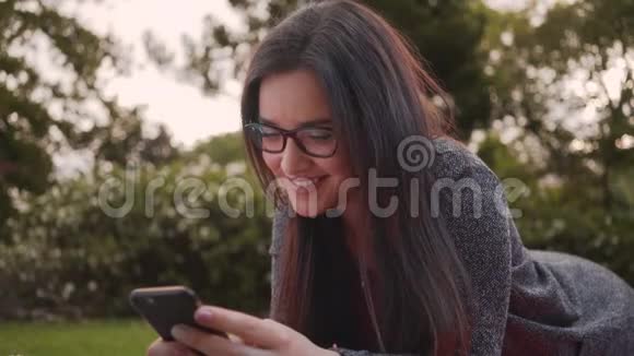 一位戴着眼镜的年轻美女躺在公园里一边看着相机一边在公园里使用智能手机视频的预览图