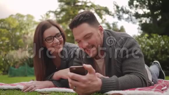 年轻夫妇躺在草地上的毯子上在公园里一起用手机上网玩得开心视频的预览图