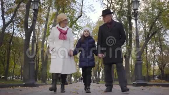 微笑的白人女孩和她的祖父母沿着公园的小巷散步穿着优雅的高级夫妇视频的预览图
