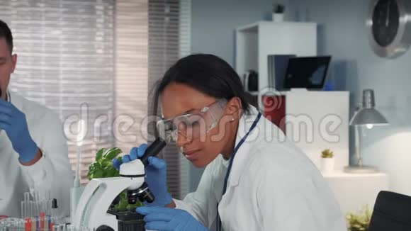 穿着实验室外套戴着安全眼镜戴着显微镜的混血女人视频的预览图