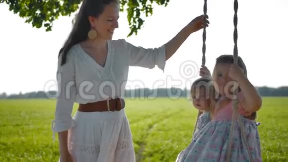 一位年轻的母亲穿着一件简单漂亮的乡村服装在秋千上摆动着她的女儿们视频的预览图