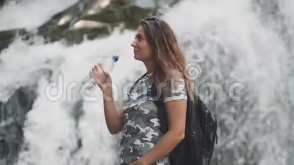 迷人的女孩从瓶子里喝冷水当站在瀑布附近的石头上时解渴侧面视频的预览图
