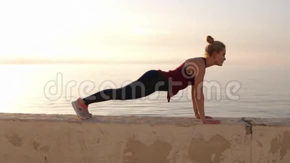 可爱的适合白种人女孩在运动腿做木板运动在海滩日落在女儿墙健身锻炼和视频的预览图