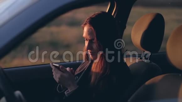 日落前的晚上坐在车里的女性使用智能手机视频的预览图
