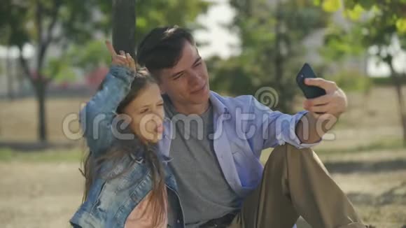 一个十几岁的男孩和他的小妹妹在智能手机上拍摄有趣的自拍照片坐在阳光明媚的秋日公园里视频的预览图