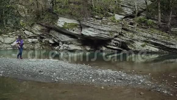 穿着紫色夹克牛仔裤和运动鞋的女孩在山上一个湖上的岩石岛上游荡视频的预览图