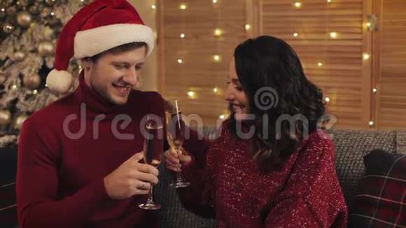 可爱的年轻夫妇坐在沙发上靠近圣诞树在舒适的家背景亲吻和举起眼镜视频的预览图