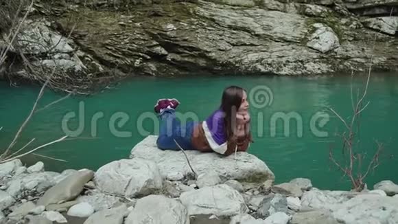 长发女孩躺在一个绿松石山湖附近的一块大石头上晃着腿视频的预览图