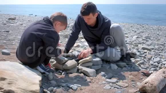 爸爸和儿子在海边的石头海滩上用木棍点燃篝火上的煤视频的预览图