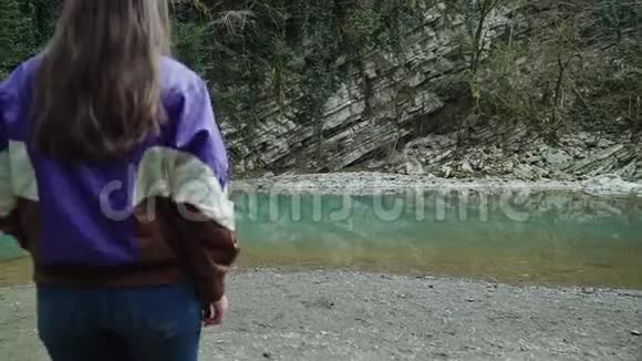 穿着夹克和牛仔裤的金发女孩来到一个清澈的蓝色山湖从后面可以看到视频的预览图