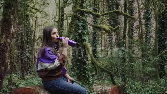 穿着运动夹克的女孩坐在森林里的树桩上她正从一个彩色的热瓶里喝酒视频的预览图