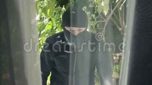 穿着黑色衣服和巴拉克拉瓦的小偷的特写镜头看着窗户准备闯入房子概念视频的预览图