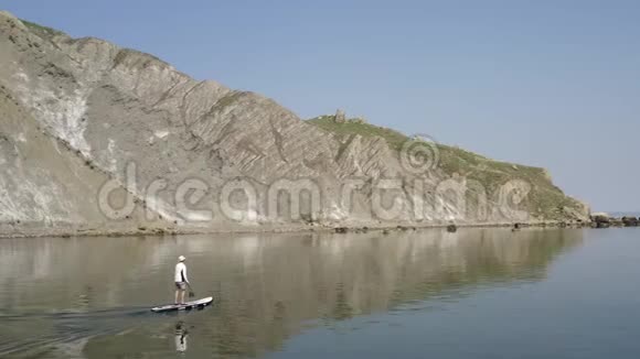孤独的冲浪者沿着平静的蓝色水沿岩石海岸慢慢移动到巨石上视频的预览图