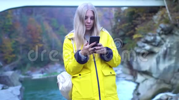 年轻迷人的金发女孩穿着黄色雨衣使用她的智能手机靠近山瀑布自然湖视频的预览图