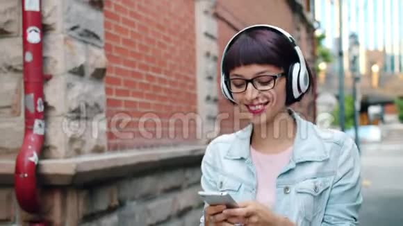 漂亮的女士用智能手机在耳机里听音乐走在街上视频的预览图