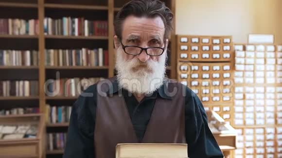 戴着眼镜的老胡子手里拿着书看着书架背景上的照相机视频的预览图