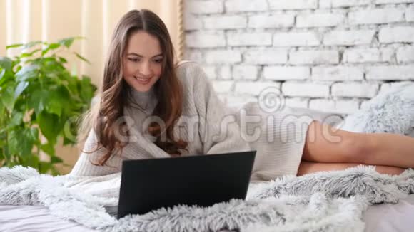 快乐的微笑美丽的女人躺在床上的笔记本电脑上聊天视频的预览图