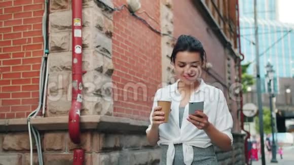 有魅力的女孩拿着智能手机拿着咖啡散步的肖像视频的预览图