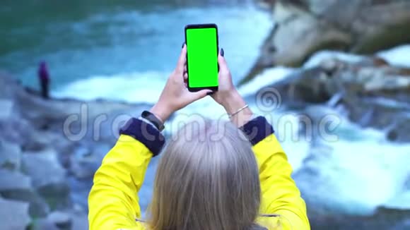 年轻的旅游女孩试图在她的智能手机上拍摄美丽的山瀑布附近的自然奇迹视频的预览图
