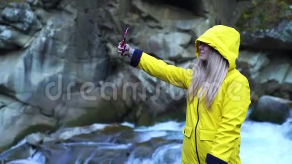 年轻迷人的金发女孩穿着黄色雨衣在瀑布上自拍在山上自然生态学生青年视频的预览图