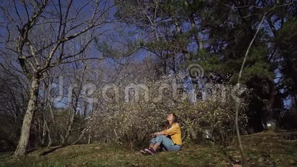 穿着牛仔裤和黄色夹克的漂亮女孩正坐在盛开的花园里的绿草上视频的预览图