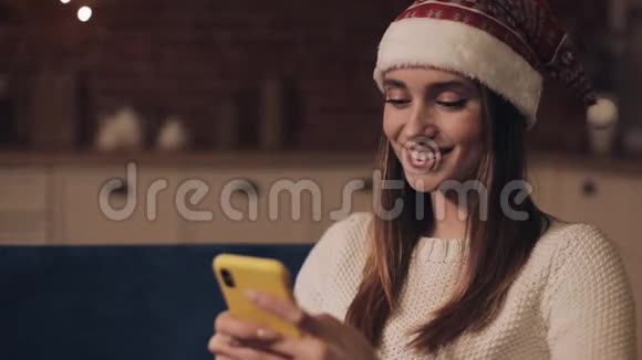 美丽微笑的年轻女孩戴着桑塔帽坐在圣诞树附近的沙发上她的智能手机上滚动着视频的预览图