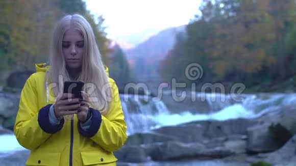 欧洲迷人的年轻女子在瀑布之间的群山中使用手机的正面景色视频的预览图