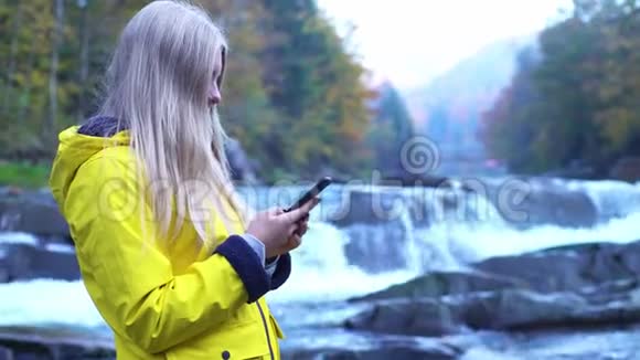 侧面图观看美丽漂亮的年轻女孩穿着黄色雨衣使用她的智能手机在瀑布附近视频的预览图