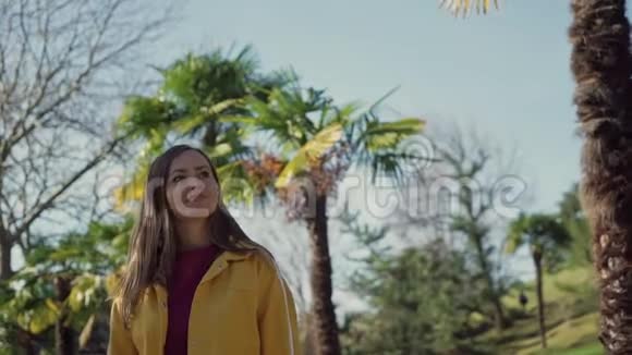 阳光明媚的一天穿着黄色夹克的美丽快乐的女孩带着棕榈树沿着大街散步视频的预览图