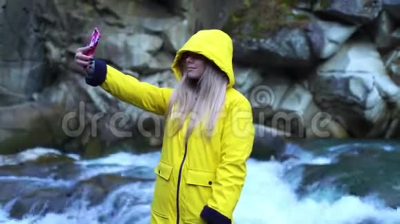 穿着黄色雨衣的漂亮女孩用智能手机在瀑布间自拍视频的预览图