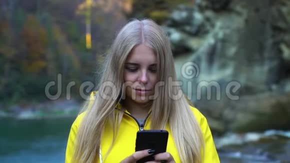 美丽迷人的白种人女孩穿着黄色雨衣用智能手机在山瀑布湖自然生态中有机结合视频的预览图