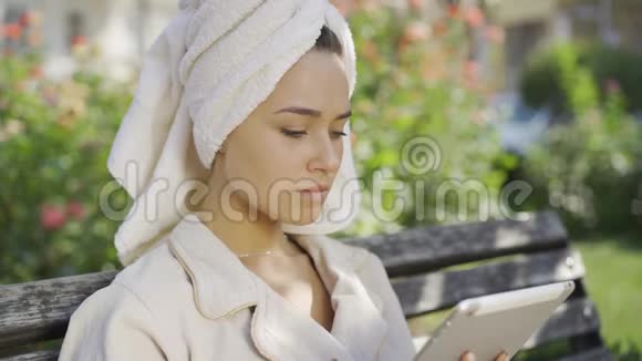公园里坐在长凳上的一位身穿浴袍头戴毛巾的年轻可爱女子的画像上面写着视频的预览图