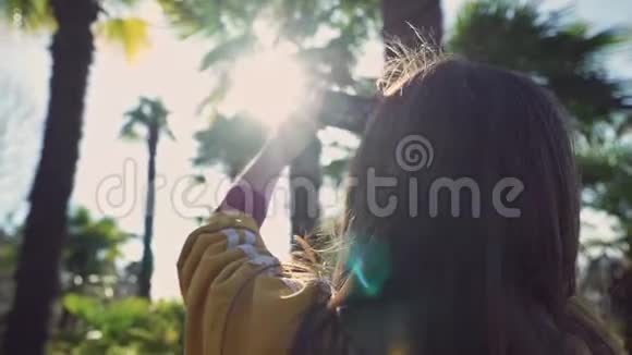 穿黄色夹克的女孩在阳光明媚的一天在特写镜头中用手机拍下棕榈树的照片视频的预览图