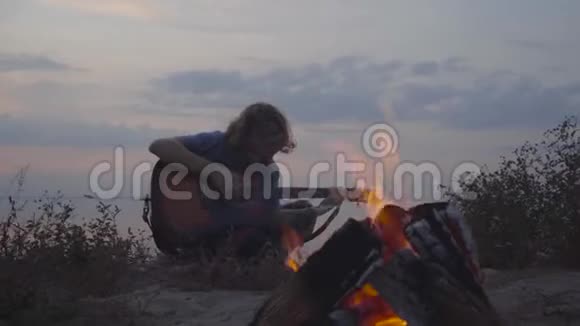 在傍晚五彩缤纷的日落和临近篝火的夏日海滩上一个年轻的小伙子在玩声吉他视频的预览图