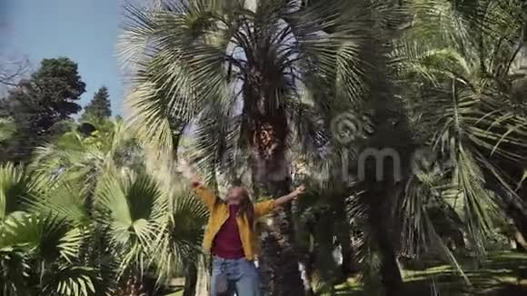 一个穿着黄色夹克衫的开朗的女孩正在玩棕榈叶在阳光明媚的日子里在棕榈树下旋转视频的预览图