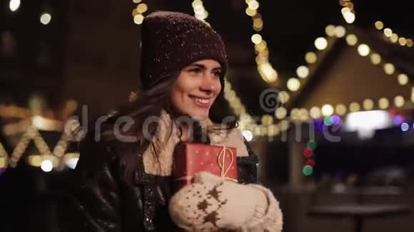 年轻可爱的微笑女孩戴着冬帽抱着礼物盒看上去又高兴又兴奋走在飘落的雪中视频的预览图