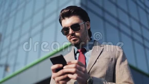 帅气的商务人士戴着太阳镜穿着时髦的领带用他的智能手机在办公室前眺望视频的预览图