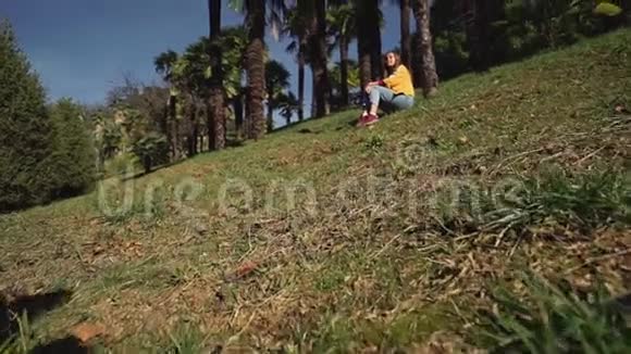 穿着牛仔裤和夹克的可爱女孩坐在棕榈树下的绿色草坪上视频的预览图