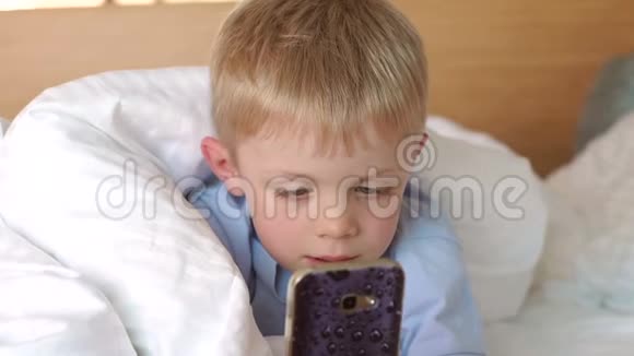 一个小男孩躺在床上躺在温暖的白色毯子下拿着一个电话视频的预览图
