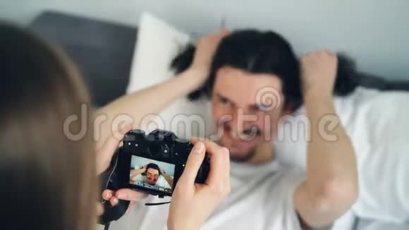 有创意的男人在床上摆着滑稽的脸而女人则用相机拍照视频的预览图
