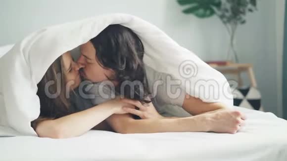 快乐的一对情侣在床上亲吻在毯子下揉鼻子一起躺在床上视频的预览图
