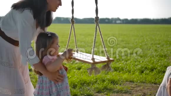 一位穿着白色连衣裙的美丽母亲帮助她的女儿坐在木制的挂绳秋千上视频的预览图