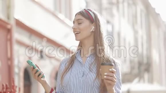 戴着时髦头巾和带条纹衬衫的幸福微笑女人戴着耳机拿着咖啡杯用她的智能手机视频的预览图