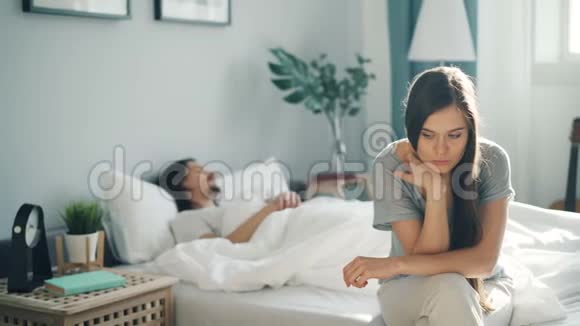 穿着睡衣的悲伤女孩坐在床上想着叹息而男人却睡在家里视频的预览图