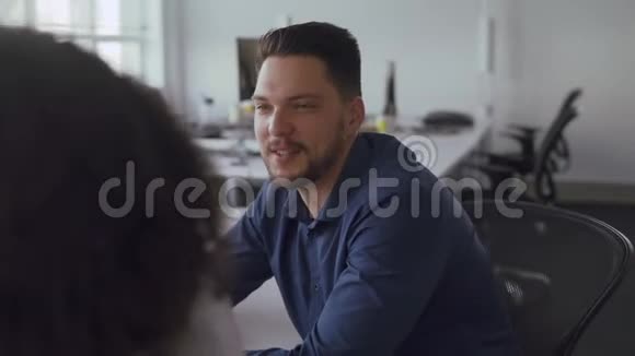 一位年轻的职业商人坐在桌子旁和一位微笑着工作的女同事讨论工作视频的预览图