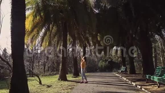 在一个阳光明媚的日子里可爱的女孩仔细检查了公园小巷上的棕榈树视频的预览图